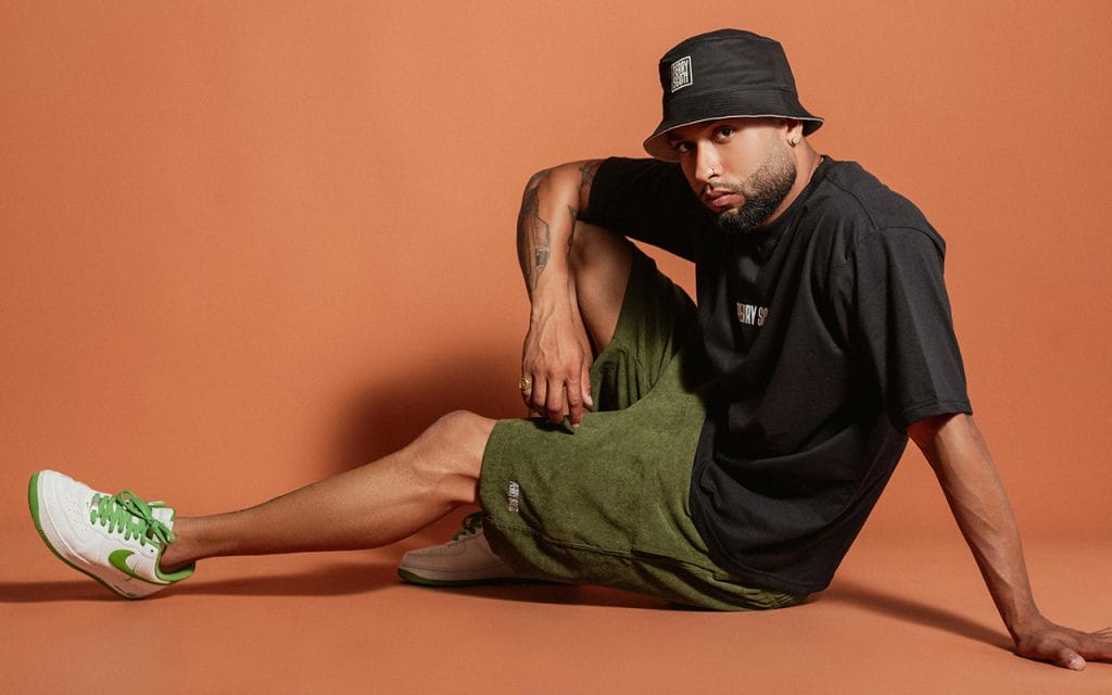 Pria berpose duduk di studio dengan latar oranye mengenakan kaus hitam, celana pendek hijau, dan sepatu putih hijau sebagai bagian dari sesi fashion photography