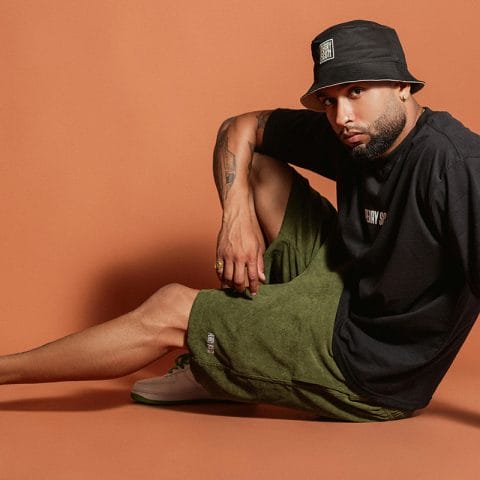 Pria berpose duduk di studio dengan latar oranye mengenakan kaus hitam, celana pendek hijau, dan sepatu putih hijau sebagai bagian dari sesi fashion photography