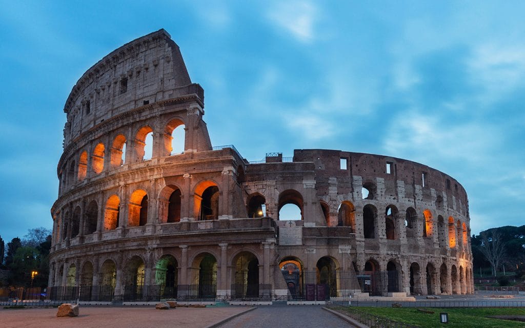 Foto arsitektur Colosseum di Roma saat senja dengan pencahayaan hangat yang menyoroti lengkungan batu kuno, menampilkan keindahan dan kemegahan arsitektur klasik Romawi