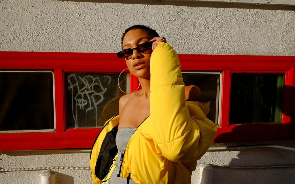 Seorang model mengenakan jaket kuning cerah dan kacamata hitam berpose di bawah sinar matahari dengan latar dinding bergaris merah, menampilkan gaya busana modern dan percaya diri