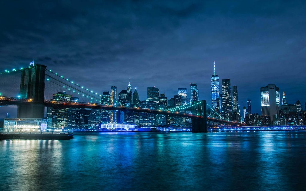 Foto kota di malam hari dengan jembatan bercahaya dan gedung tinggi berlampu, menampilkan teknik fotografi malam hari