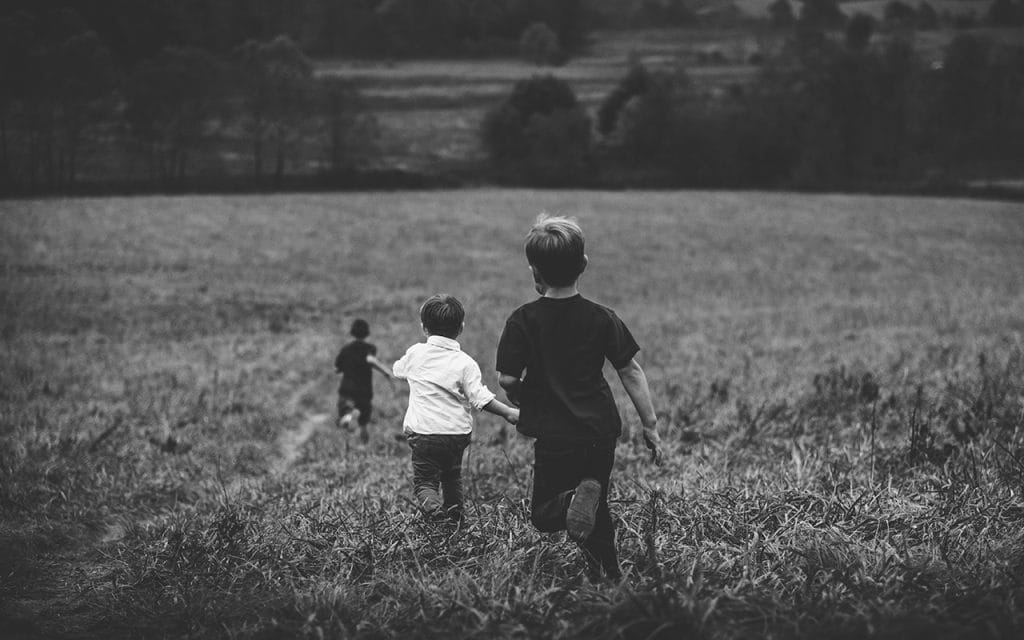 Anak-anak berlari melintasi padang rumput dalam foto hitam putih yang menonjolkan suasana nostalgia dan kebebasan masa kecil