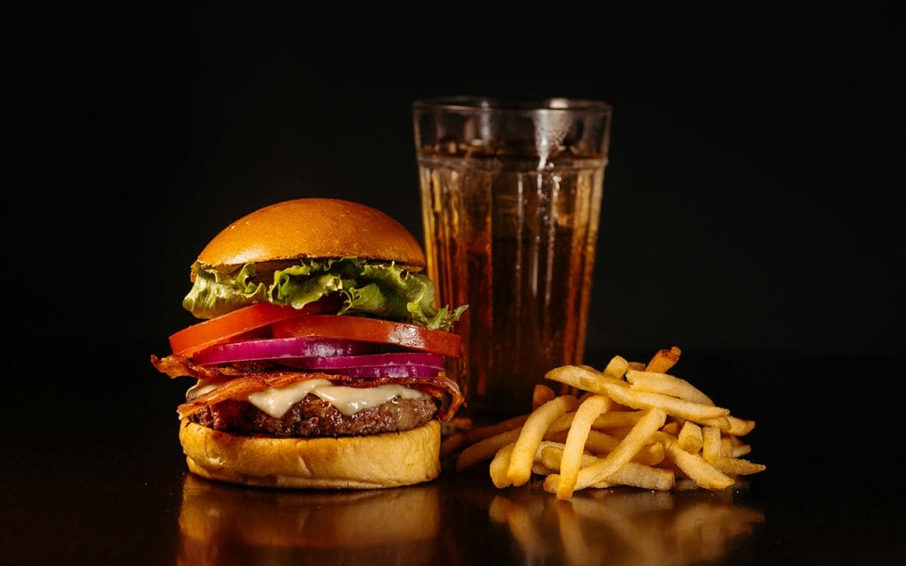 Foto close-up burger dengan keju, tomat, bawang, dan selada disajikan bersama kentang goreng dan segelas soda di atas meja gelap, menggambarkan konsep fotografi makanan dengan pencahayaan dramatis dan detail yang menggugah selera