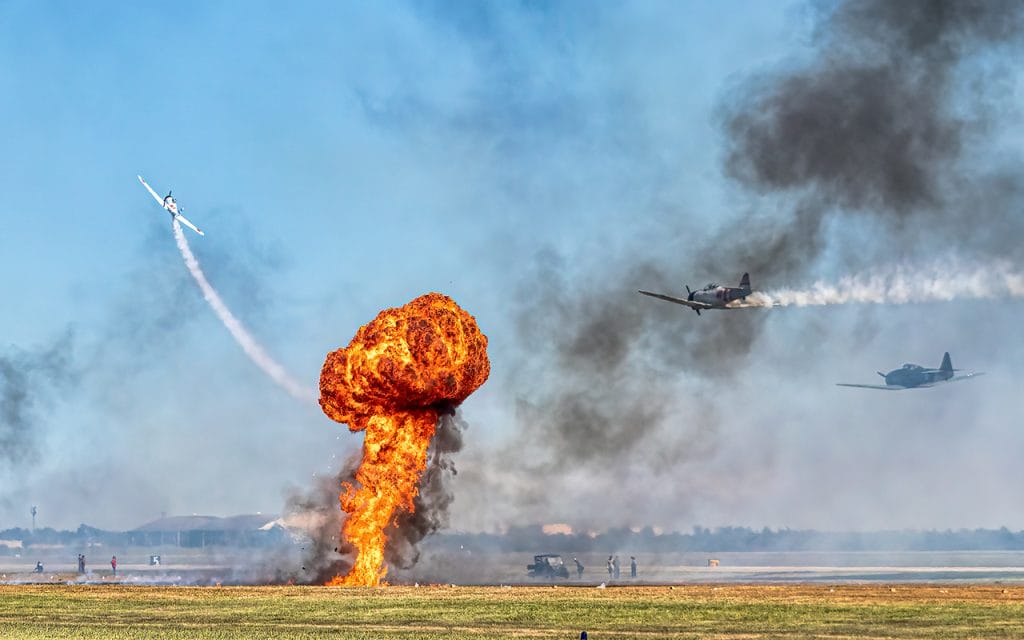 Foto pesawat tempur terbang di langit dengan ledakan besar di landasan, menggambarkan intensitas dan dramatisnya suasana dalam fotografi peperangan