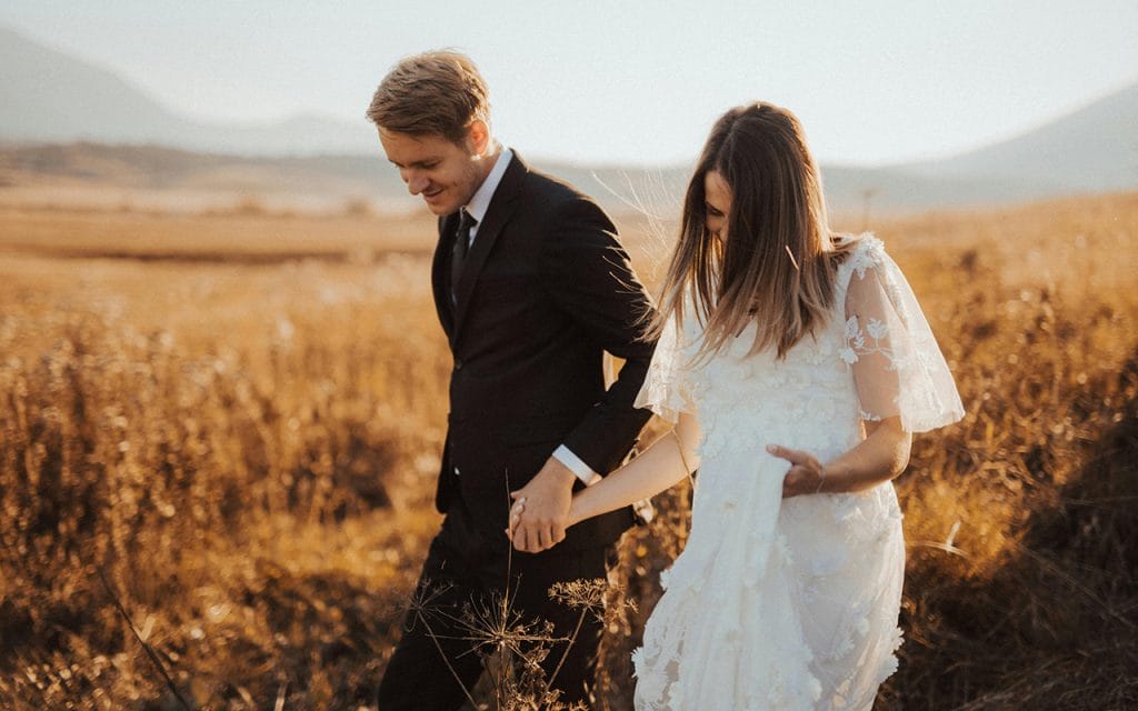 Pasangan pengantin berjalan di ladang saat matahari terbenam, hasil pemotretan fotografi pernikahan bergaya alami dan romantis