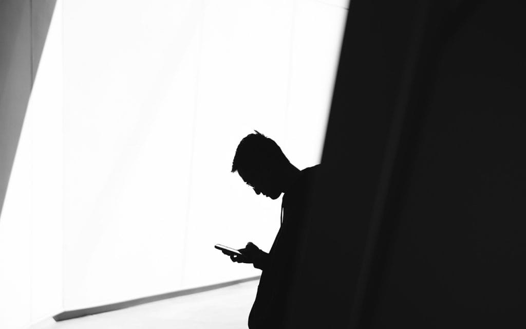 Siluet seseorang sedang melihat ponsel di area minim cahaya dengan latar terang, menggambarkan konsep fotografi rahasia yang menangkap momen diam-diam penuh misteri