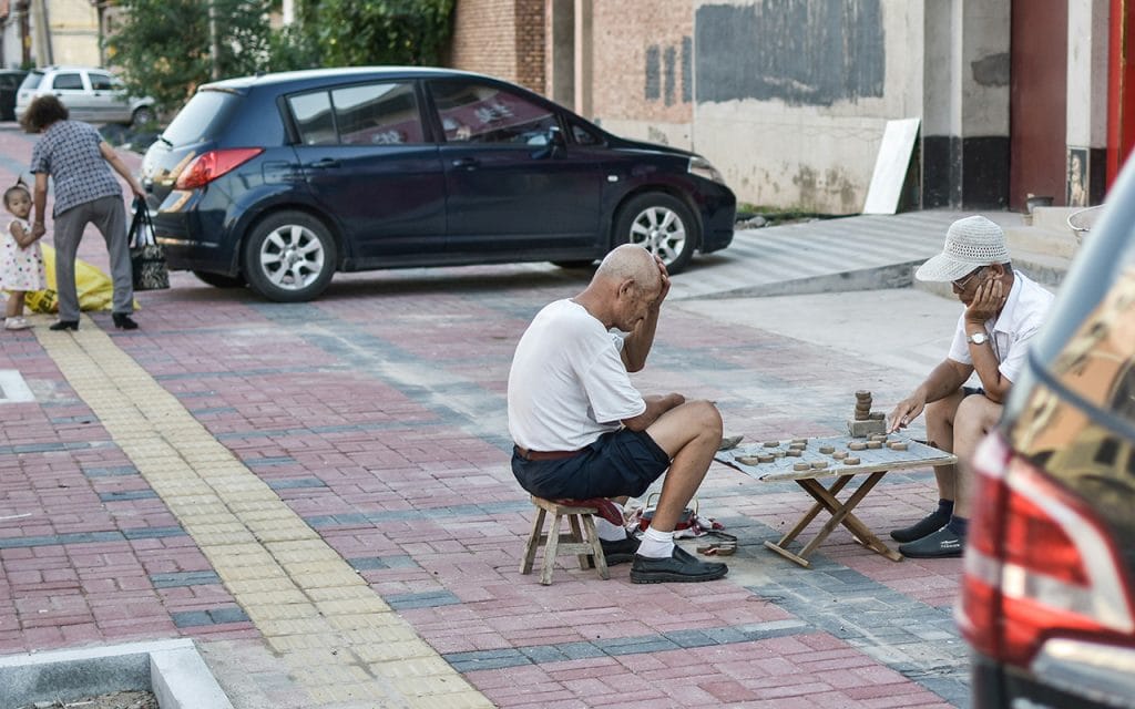 Dua pria lanjut usia duduk di pinggir jalan sambil bermain papan permainan sederhana, menggambarkan momen keseharian yang spontan dan alami dalam gaya fotografi vernacular