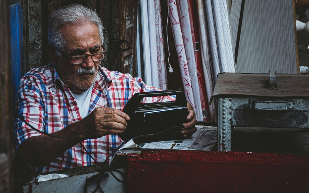 Seorang pria tua mengenakan kemeja kotak-kotak sedang memperbaiki radio di bengkel kecil, contoh candid photography yang natural