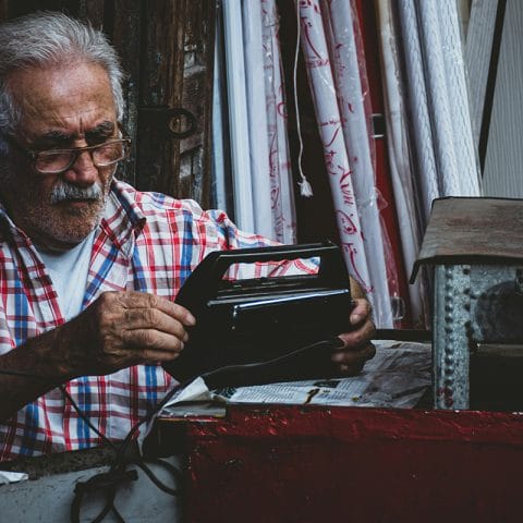 Seorang pria tua mengenakan kemeja kotak-kotak sedang memperbaiki radio di bengkel kecil, contoh candid photography yang natural