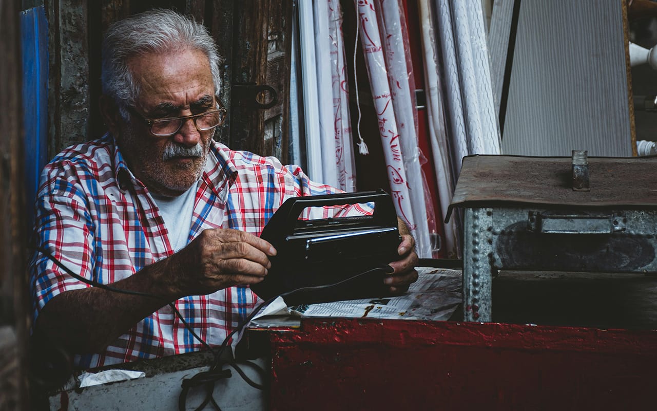 Seorang pria tua mengenakan kemeja kotak-kotak sedang memperbaiki radio di bengkel kecil, contoh candid photography yang natural