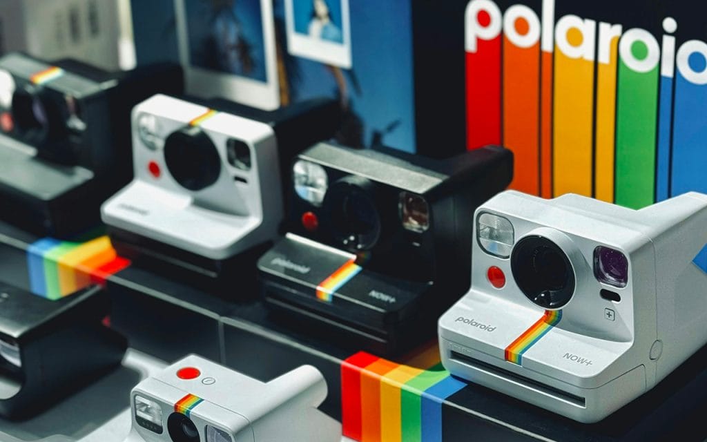Deretan kamera polaroid berbagai model warna hitam dan putih dipajang di etalase toko dengan latar logo Polaroid berwarna pelangi