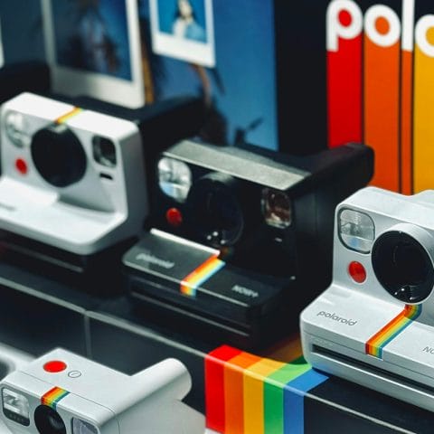 Deretan kamera polaroid berbagai model warna hitam dan putih dipajang di etalase toko dengan latar logo Polaroid berwarna pelangi