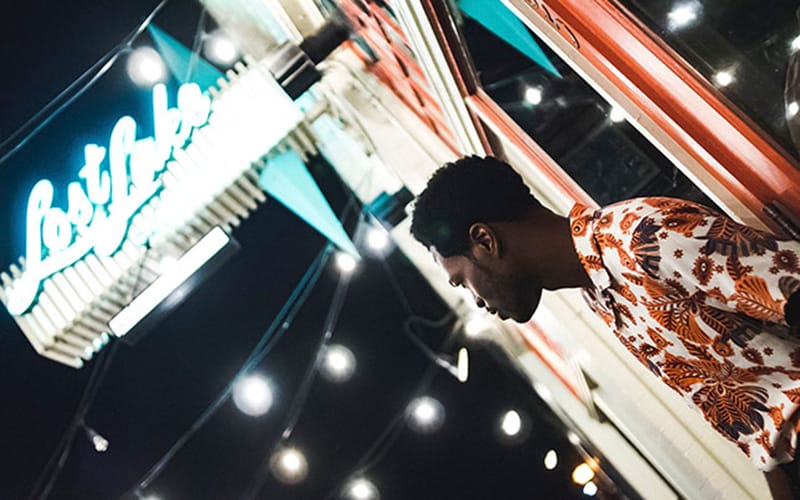 Foto sudut Canted Angle menampilkan seorang pria berdiri di depan bangunan bercahaya malam hari, dengan kamera dimiringkan untuk menciptakan kesan dramatis dan perspektif yang unik