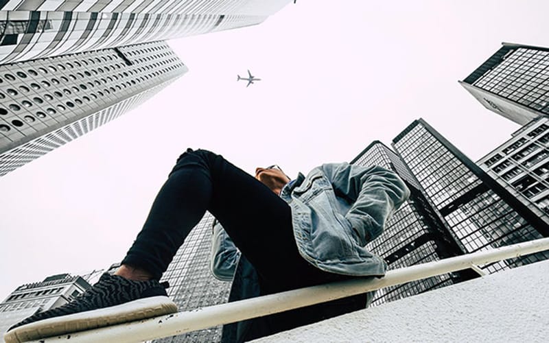 Foto sudut Frog Eye View menampilkan seseorang berpose di antara gedung-gedung tinggi dengan pesawat terbang melintas di langit, diambil dari sudut rendah untuk menciptakan kesan dramatis dan megah
