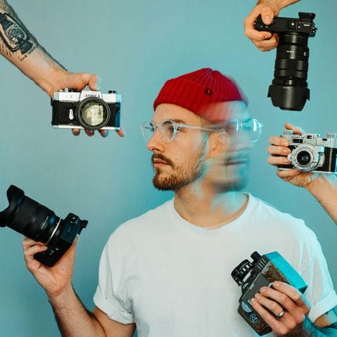 Seorang pria memakai topi merah dan kacamata dikelilingi beberapa kamera yang diarahkan padanya, melambangkan berbagai sudut pengambilan gambar dalam fotografi